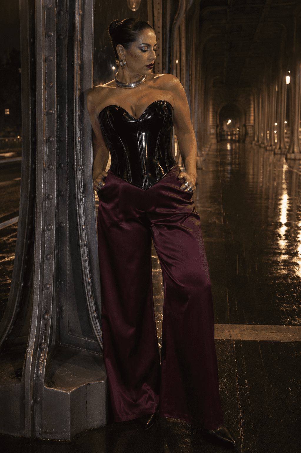 Bustier corset gothique moderne noir vernis porté avec un pantalon fluide satin bordeaux au Pont Bir-Hakeim à Paris lors d’une séance photo éditoriale de nuit sous une pluie fine.