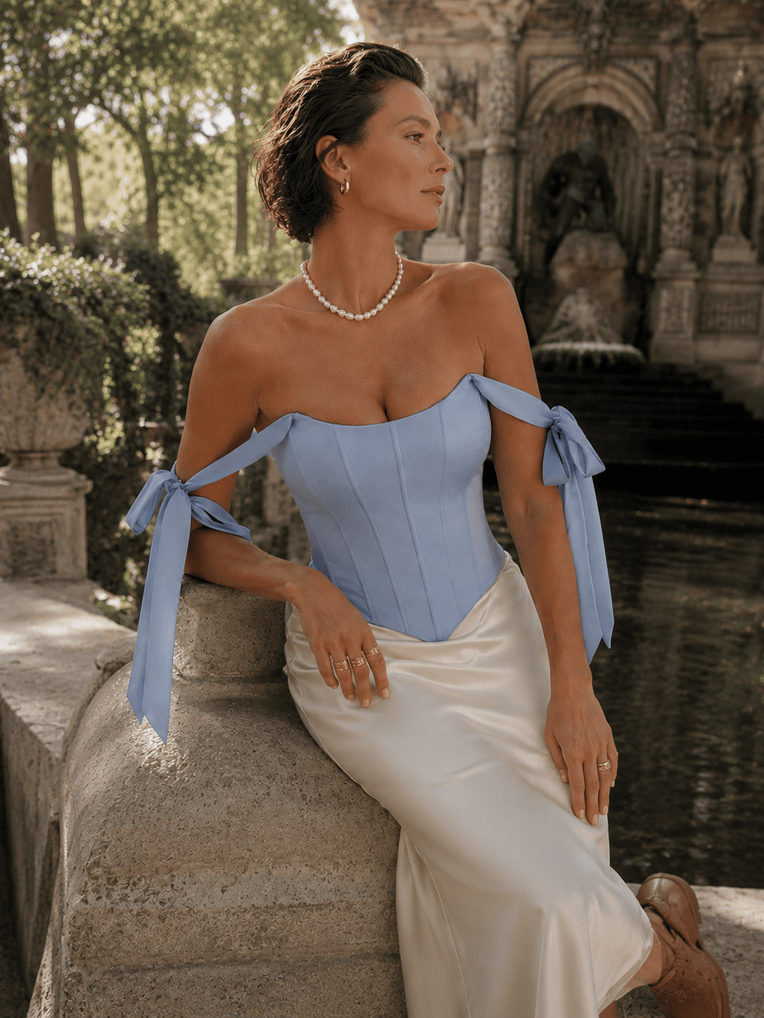 Femme de 40 ans portant un top corset satiné bleu pastel avec jupe midi satinée écrue au Jardin du Luxembourg devant la fontaine Médicis à Paris.