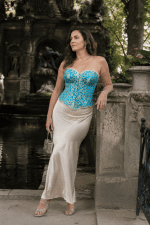 Femme de 45 ans portant un bustier corset court turquoise et or à motifs baroques avec une longue jupe satinée crème au bord de la fontaine Médicis du Jardin du Luxembourg.