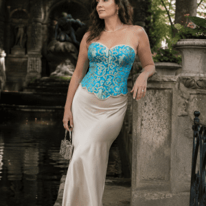 Femme de 45 ans portant un bustier corset court turquoise et or à motifs baroques avec une longue jupe satinée crème au bord de la fontaine Médicis du Jardin du Luxembourg.