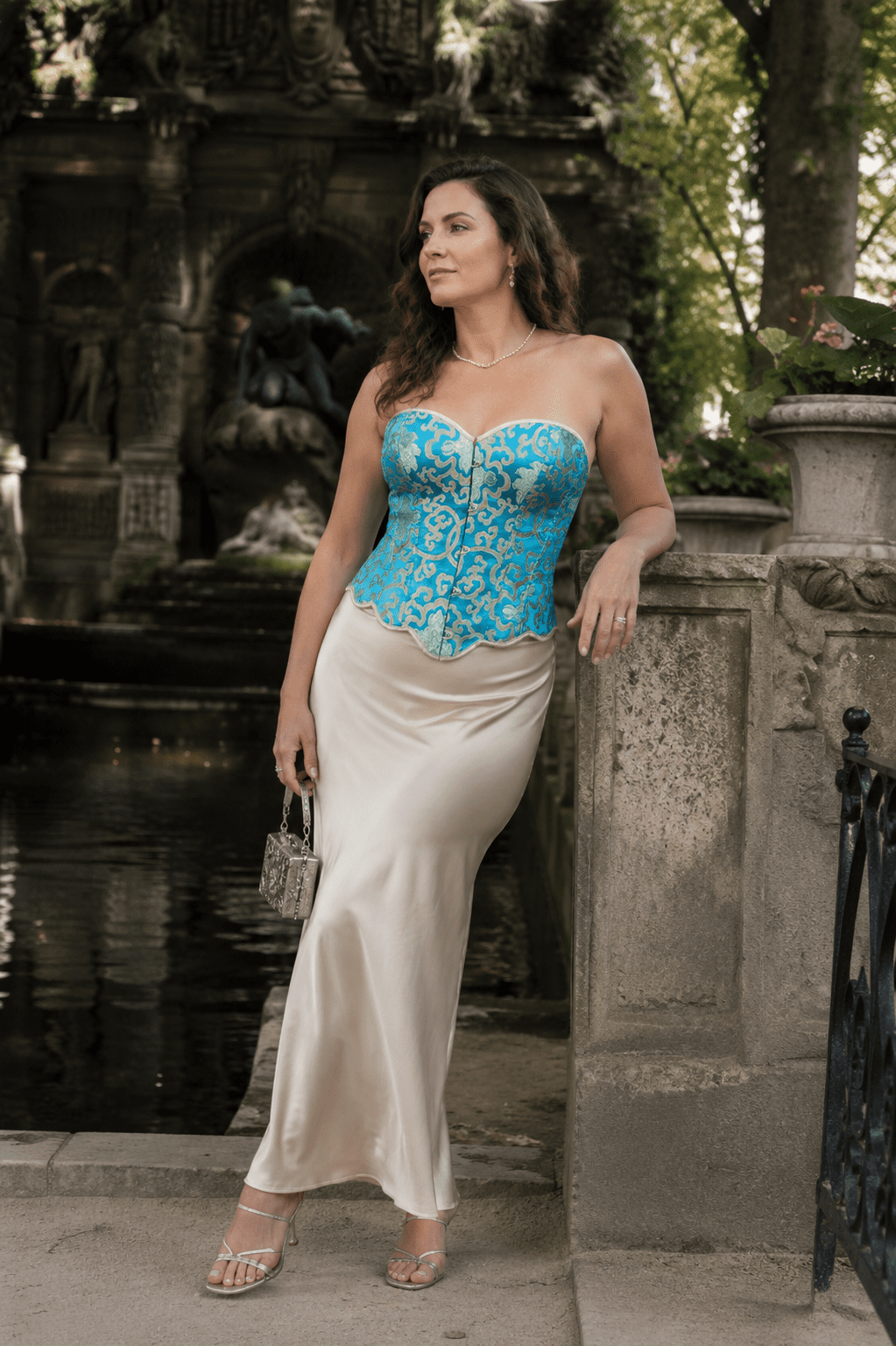 Femme de 45 ans portant un bustier corset court turquoise et or à motifs baroques avec une longue jupe satinée crème au bord de la fontaine Médicis du Jardin du Luxembourg.