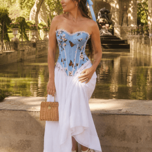Femme de 40 ans portant un corset bleu ciel imprimé de papillons et fleurs avec finitions en dentelle blanche, associé à une longue jupe fluide blanche à la fontaine de Médicis à Paris durant la golden hour du matin.