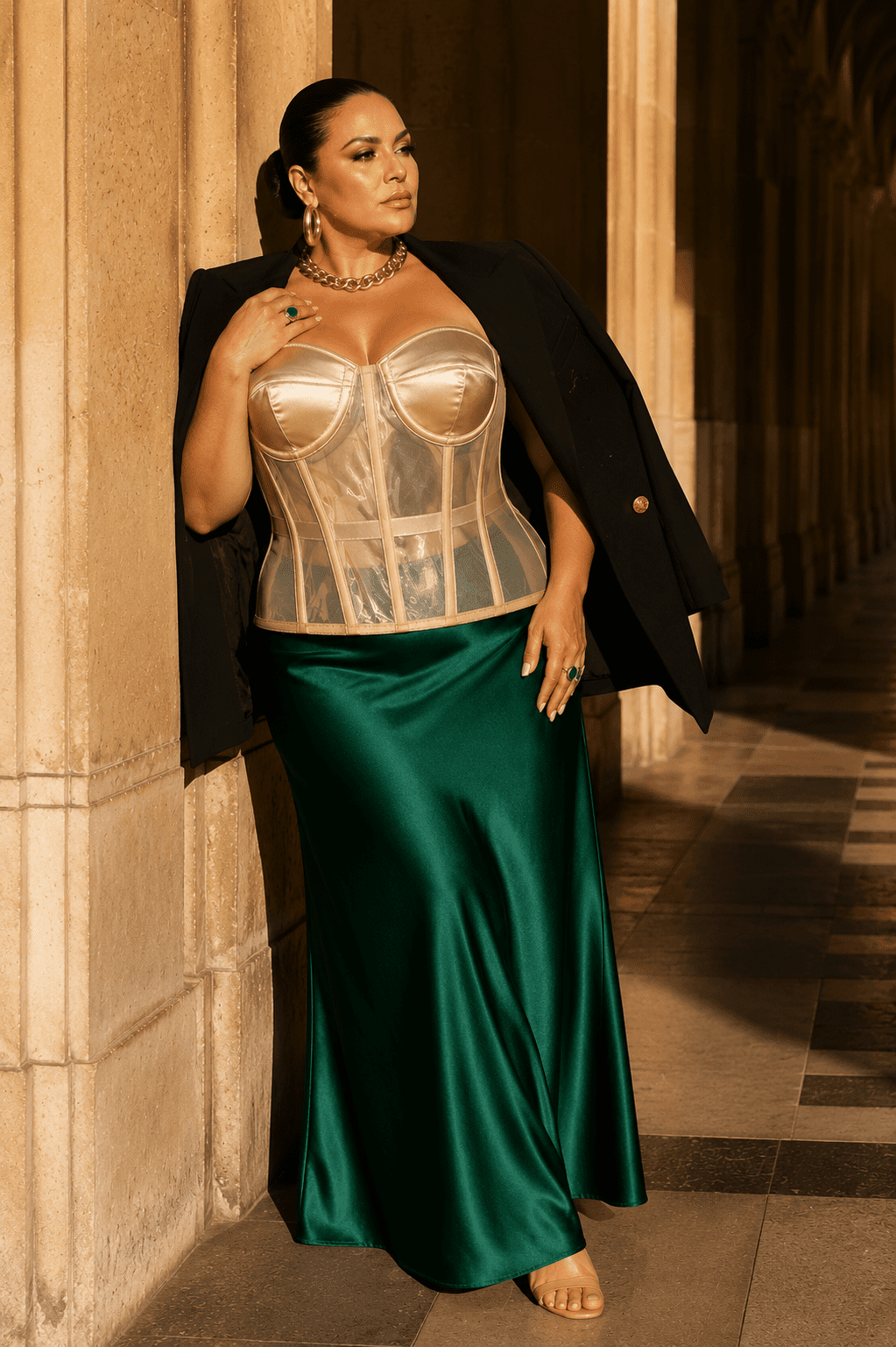 Femme de 40 ans taille 3XL portant un corset bustier transparent beige grande taille avec bonnets satinés champagne, une jupe longue satinée vert émeraude et un blazer noir oversize posé sur les épaules, appuyée contre une colonne de la Galerie Valois au Palais Royal dans une lumière dorée de fin d'après-midi.