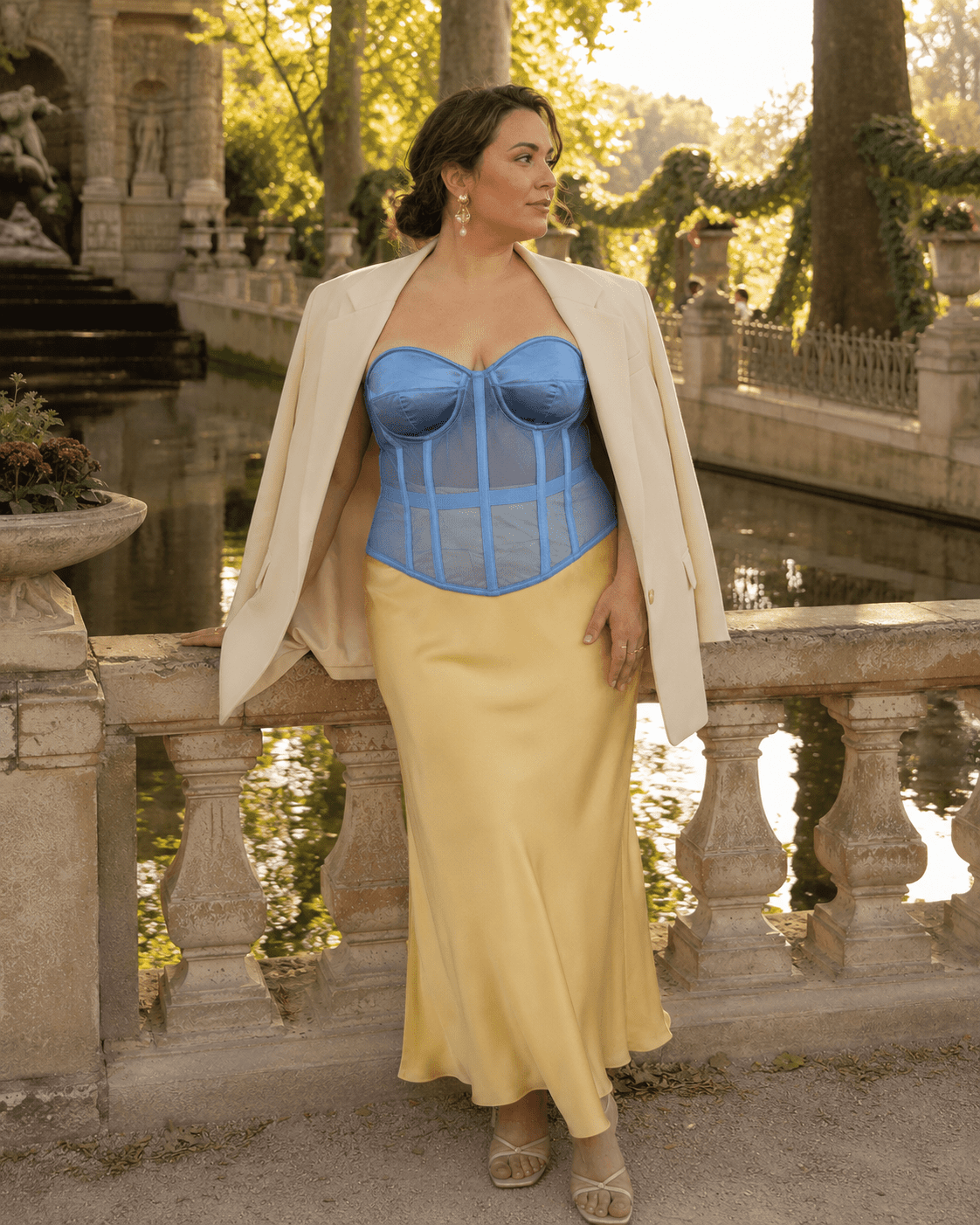 Femme de 40 ans taille 3XL portant un corset bustier bleu transparent grande taille, une jupe satin jaune beurre et un blazer crème posé sur les épaules à la fontaine de Médicis.