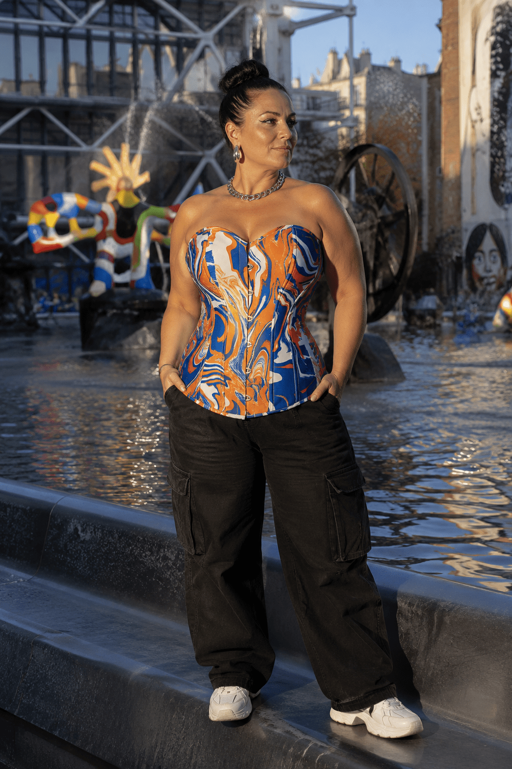 Femme de 45 ans portant un corset bustier psychédélique bleu roi, orange et blanc avec un pantalon cargo noir sur la place Igor-Stravinsky à Paris, devant la fontaine Stravinsky au coucher du soleil.