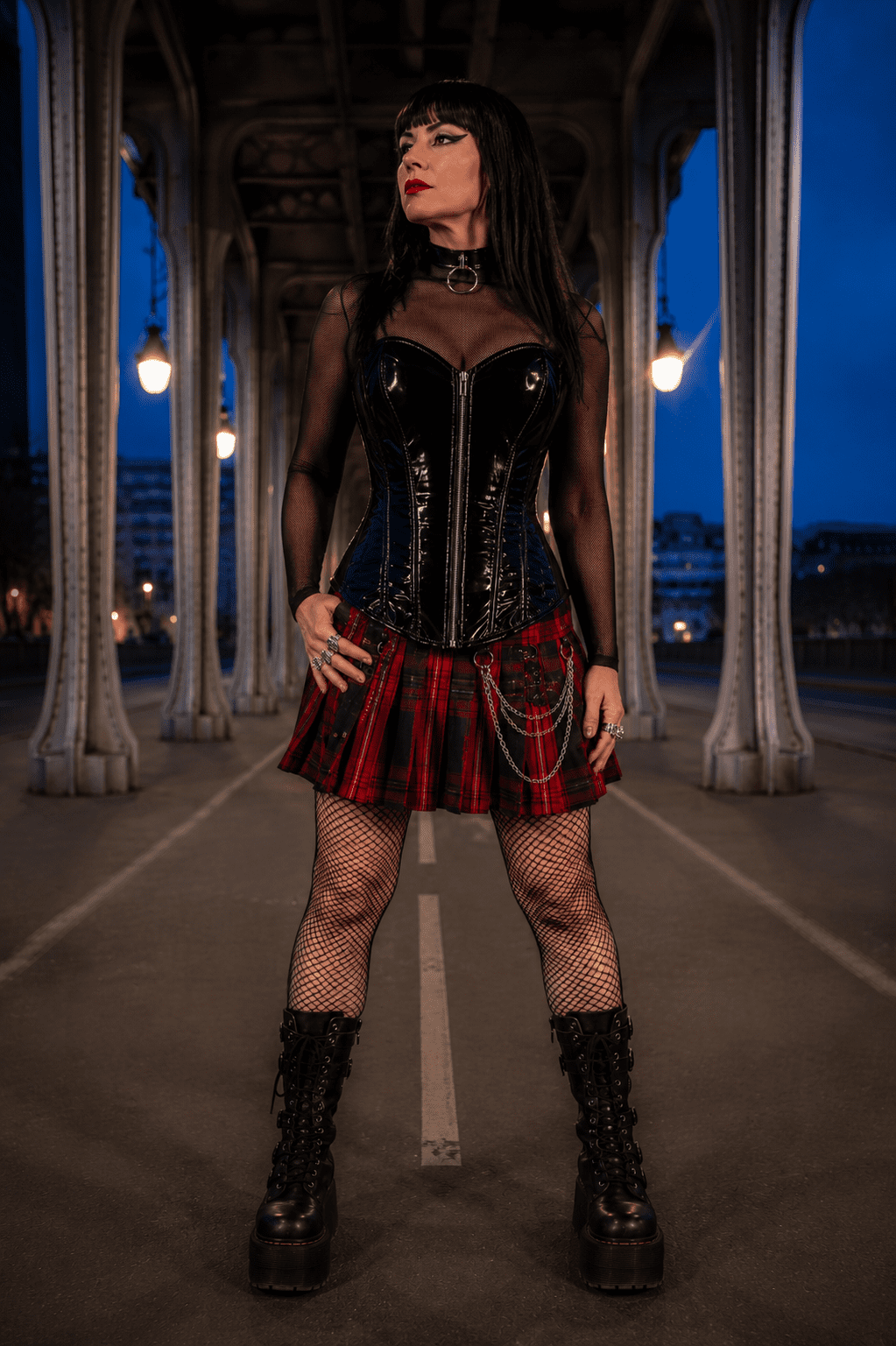 Corset bustier noir en latex brillant porté sur un top en résille noire à manches longues avec minijupe écossaise rouge et noire au pont Bir-Hakeim à Paris à la blue hour.