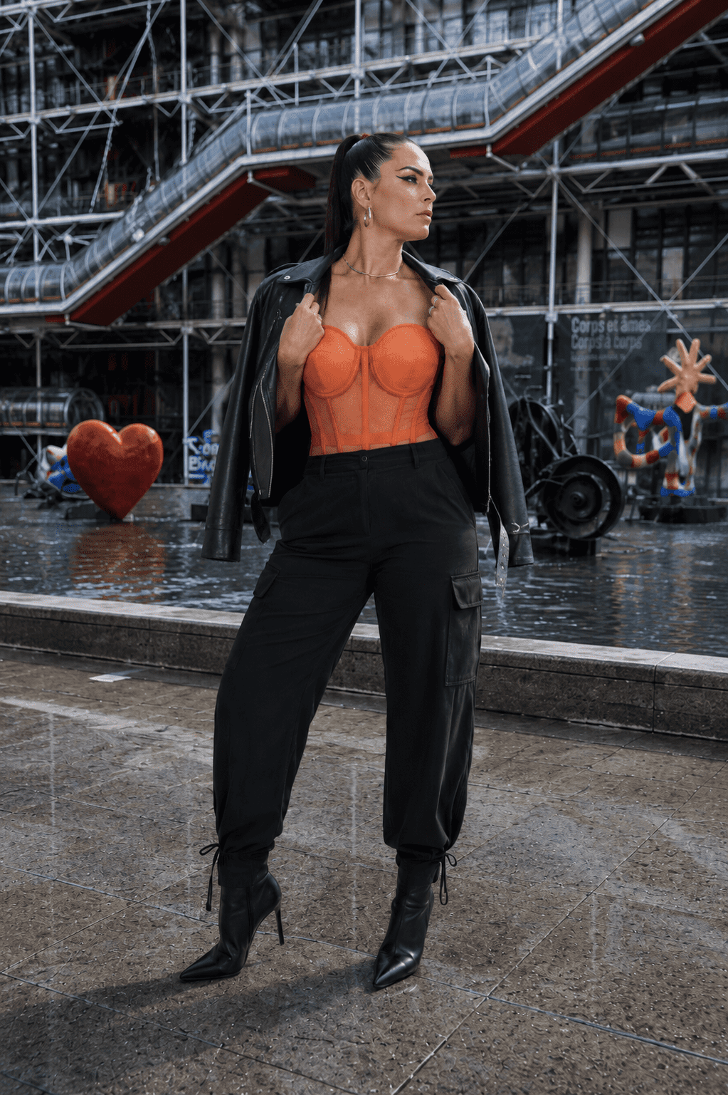 Femme portant un corset bustier transparent orange avec un perfecto noir posé sur les épaules et un pantalon cargo noir à la place Igor Stravinsky à Paris.
