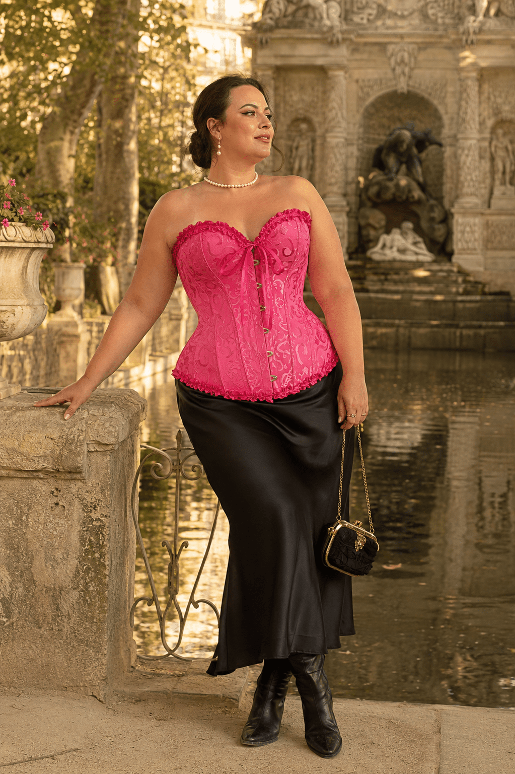 Femme de 40 ans portant un corset bustier victorien rose grande taille avec motif jacquard ton sur ton, jupe midi noire en satin et bottines victoriennes à la Fontaine de Médicis à Paris.