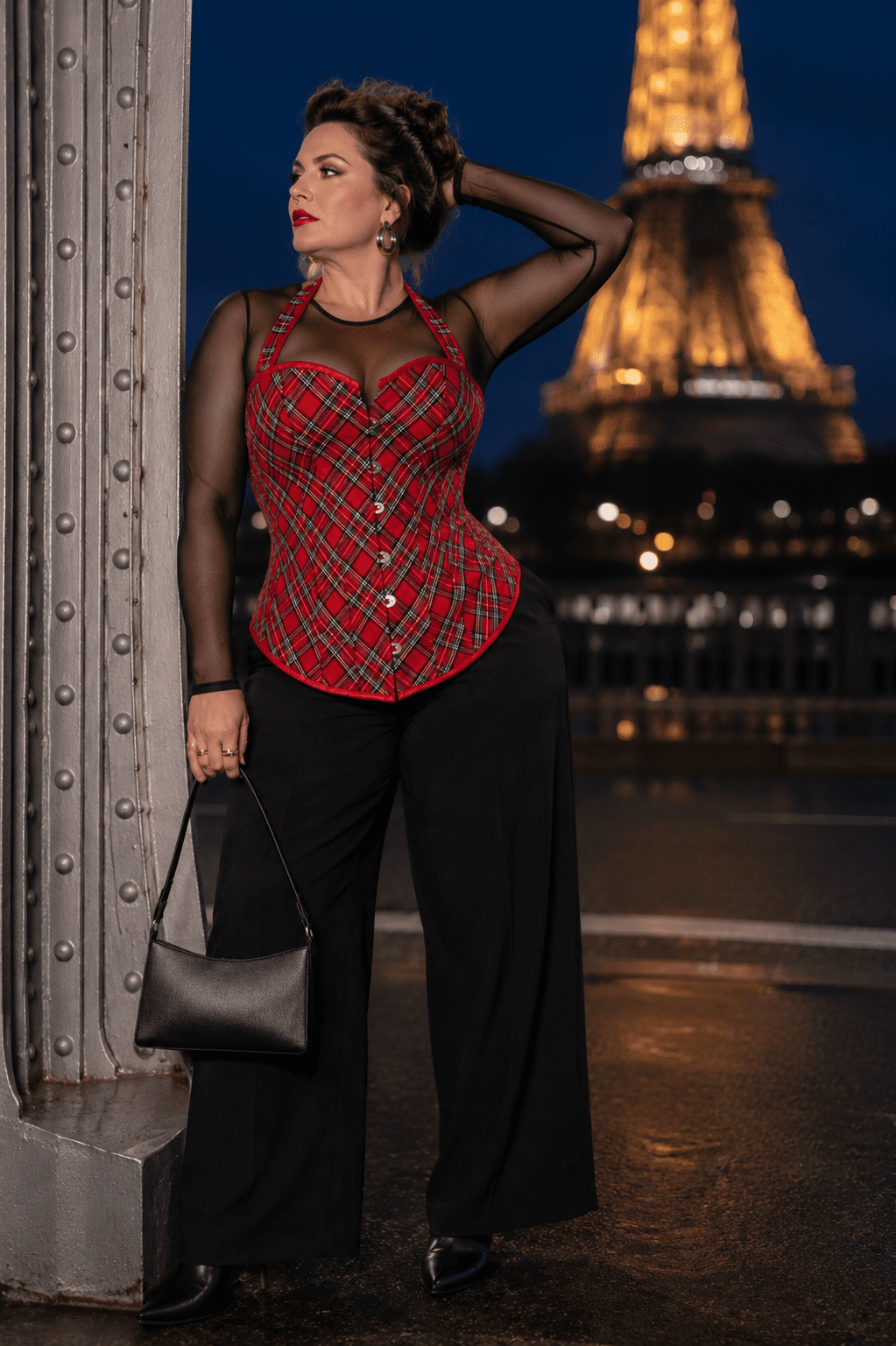 Photo de mode éditoriale au Pont Bir-Hakeim à Paris à la blue hour mettant en scène un corset bustier écossais rouge et gris à motif tartan porté sur un body en tulle noir transparent à manches longues. Le corset est associé à un pantalon de tailleur noir fluide, des bottines pointues en cuir noir et un sac baguette noir, avec la tour Eiffel illuminée et floutée en arrière-plan.