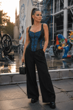 Photo éditoriale de mode d'un corset gothique romantique néo-victorien bleu roi en dentelle avec empiècements en similicuir noir, porté avec un pantalon noir fluide taille haute sur la place Stravinsky à Paris.