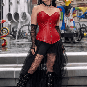 Femme de 40 ans portant un corset gothique rouge rubis en simili-cuir avec détails métalliques, jupe asymétrique en tulle noir et bottes plateforme style cybergoth à la place Igor-Stravinsky à Paris.