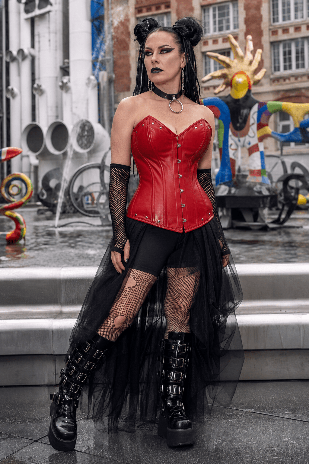 Femme de 40 ans portant un corset gothique rouge rubis en simili-cuir avec détails métalliques, jupe asymétrique en tulle noir et bottes plateforme style cybergoth à la place Igor-Stravinsky à Paris.