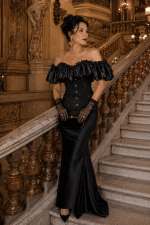 Photo de mode éditoriale au Palais Garnier à Paris mettant en scène un corset bustier noir victorien à manches bouffantes porté avec une longue jupe sirène en satin noir. Le look est complété par des gants en tulle noir, des bijoux dorés baroques et une pochette en velours noir, dans l'ambiance raffinée du grand escalier de marbre.
