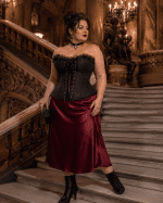 Femme de 40 ans taille 3XL portant un corset noir néo-victorien grande taille avec jupe midi satin bordeaux dans le grand escalier du Palais Garnier à Paris.