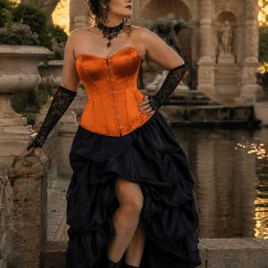 Femme de 45 ans portant un corset orange écarlate satiné néo-victorien avec jupe asymétrique noire en taffetas à la Fontaine de Médicis à Paris durant la golden hour du matin.