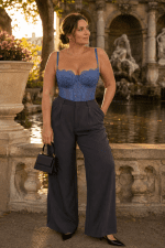 Photo éditoriale de mode réalisée à la fontaine Médicis à Paris durant la golden hour du matin. Le modèle de 40 ans porte un corset soutien-gorge bleu pastel en dentelle florale semi-transparente avec bretelles fines, associé à un pantalon de tailleur large gris anthracite taille haute. La silhouette élégante est complétée par des slingbacks noirs à bout pointu et des bijoux dorés raffinés.