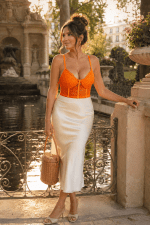 Femme de 40 ans portant un corset soutien-gorge orange en dentelle avec une jupe midi satin crème à la fontaine de Médicis à Paris durant la golden hour du matin.