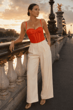 Femme de 40 ans portant un corset soutien-gorge rouge corail en dentelle avec armatures et un pantalon de tailleur crème taille haute au Pont Alexandre III à Paris durant la golden hour.