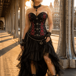 Photo de mode éditoriale au Pont Bir-Hakeim à Paris mettant en scène un corset steampunk victorien noir et rouge à panneaux contrastés et laçages décoratifs, porté avec une jupe asymétrique en tulle noir, des bottines victoriennes et des accessoires gothiques raffinés dans une lumière dorée de fin d’après-midi.