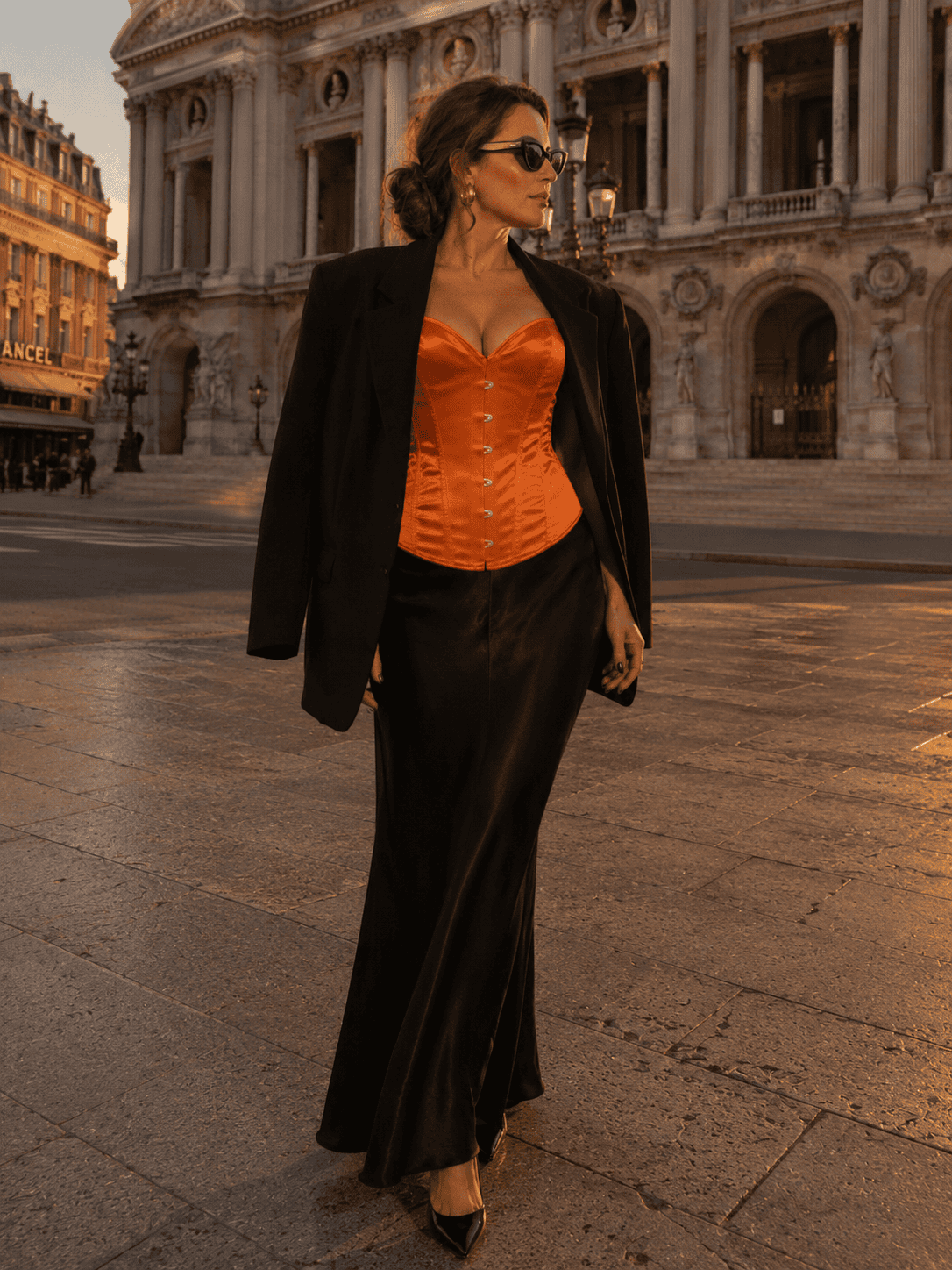 Femme élégante portant un corset bustier satin orange brûlé avec blazer noir oversize et lunettes œil de chat sur le parvis du palais garnier.