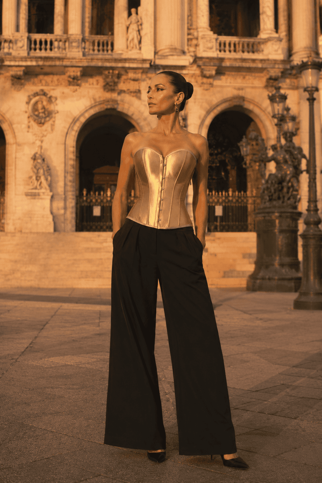 Corset bustier steampunk doré métallique porté avec un pantalon palazzo noir sur le parvis du palais garnier à Paris au coucher du soleil.