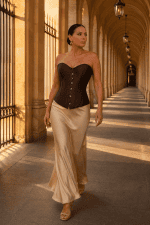 Femme de 45 ans portant un corset satin marron gothique neo-victorien avec une jupe satinée crème et un long trench beige au Pont Alexandre III à Paris à la golden hour.