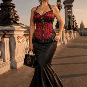 Femme de 45 ans portant un corset steampunk en brocart rouge carmin et similicuir noir avec jupe sirène noire en satin duchesse au Pont Alexandre III à Paris, à la golden hour du soir.