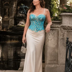 Femme de 45 ans portant un bustier corset court turquoise et or à motifs baroques avec une longue jupe satinée crème au bord de la fontaine Médicis du Jardin du Luxembourg.
